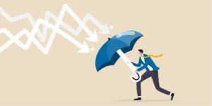 The image depicts a person holding a large umbrella, shielding themselves from descending arrows representing declining graphs or data trends. The arrows symbolize a downturn or negative trend, while the umbrella suggests protection and resilience against these adverse conditions. The individual, dressed in business attire, seems to be bracing themselves, indicating a proactive stance against challenges. The image conveys themes of crisis management, risk mitigation, resilience in the face of business challenges, and strategies to safeguard against negative impacts.