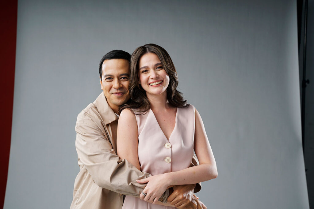 Filipino love teams Bea Alonzo and John Lloyd Cruz in a studio portrait, wearing coordinating neutral tones with Bea in a soft pink sleeveless top and John Lloyd in a beige shirt, smiling warmly together against a minimalist gray background