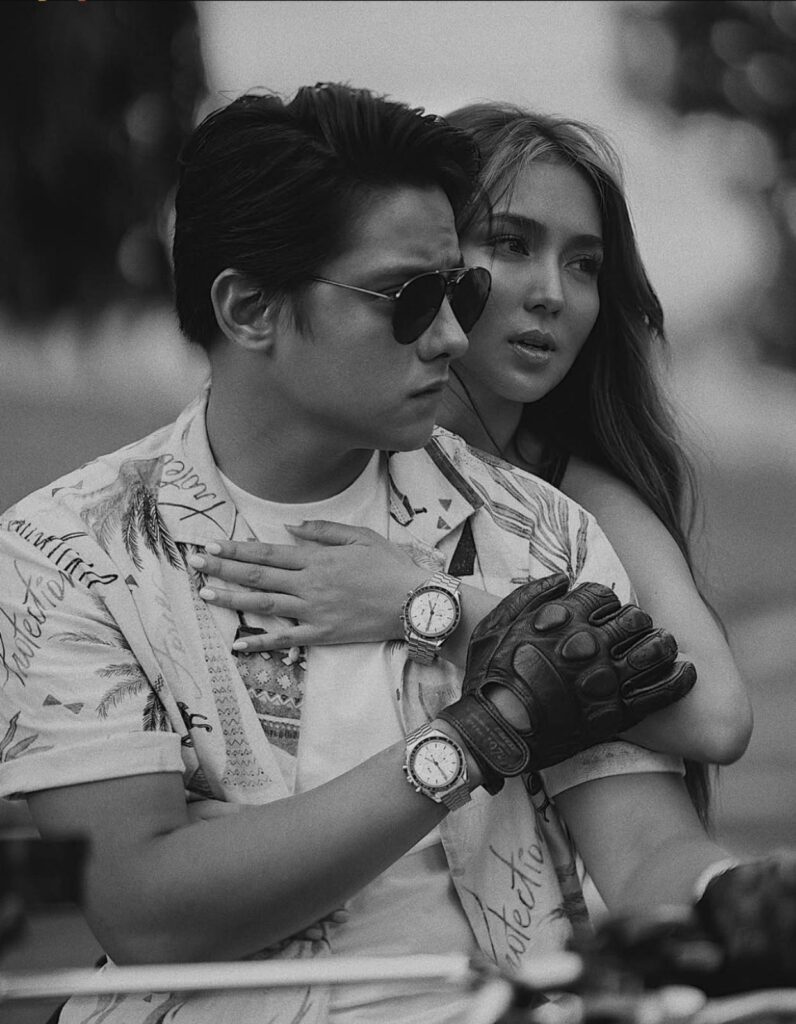 Filipino love teams Kathryn Bernardo and Daniel Padilla in black and white photography, wearing matching watches and casual tropical print shirts at an outdoor event